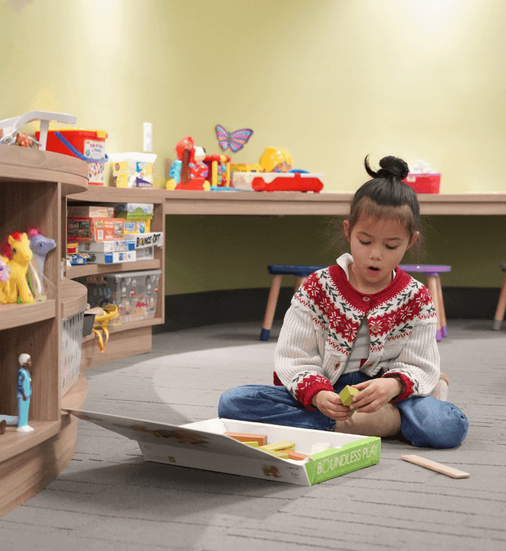 Child playing with wooden blocks on the floor by curved shelves filled with colorful toys and games at Hotel X Toronto