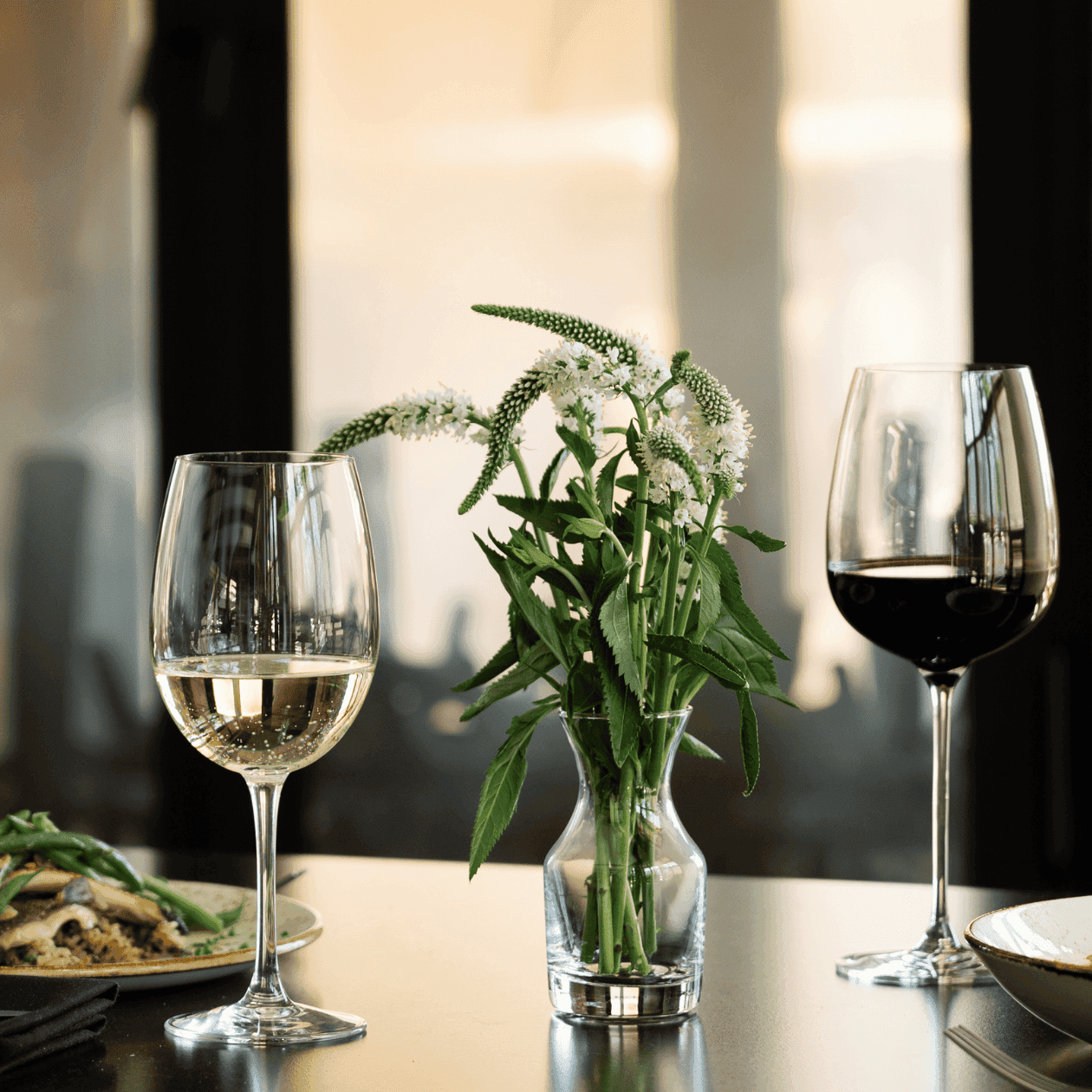Two wine glasses, a flower vase, and two plates of food on a table for Valentine's Dinner 2026.