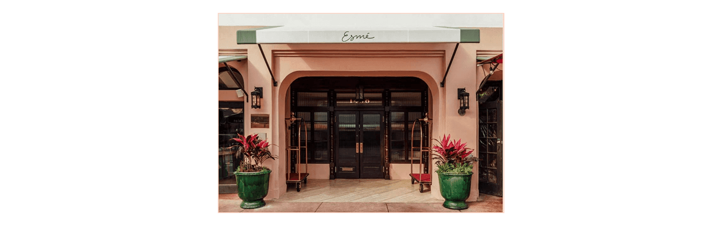 The exterior of a hotel with a pink facade, arched entryway, and green planters on either side of the entrance.