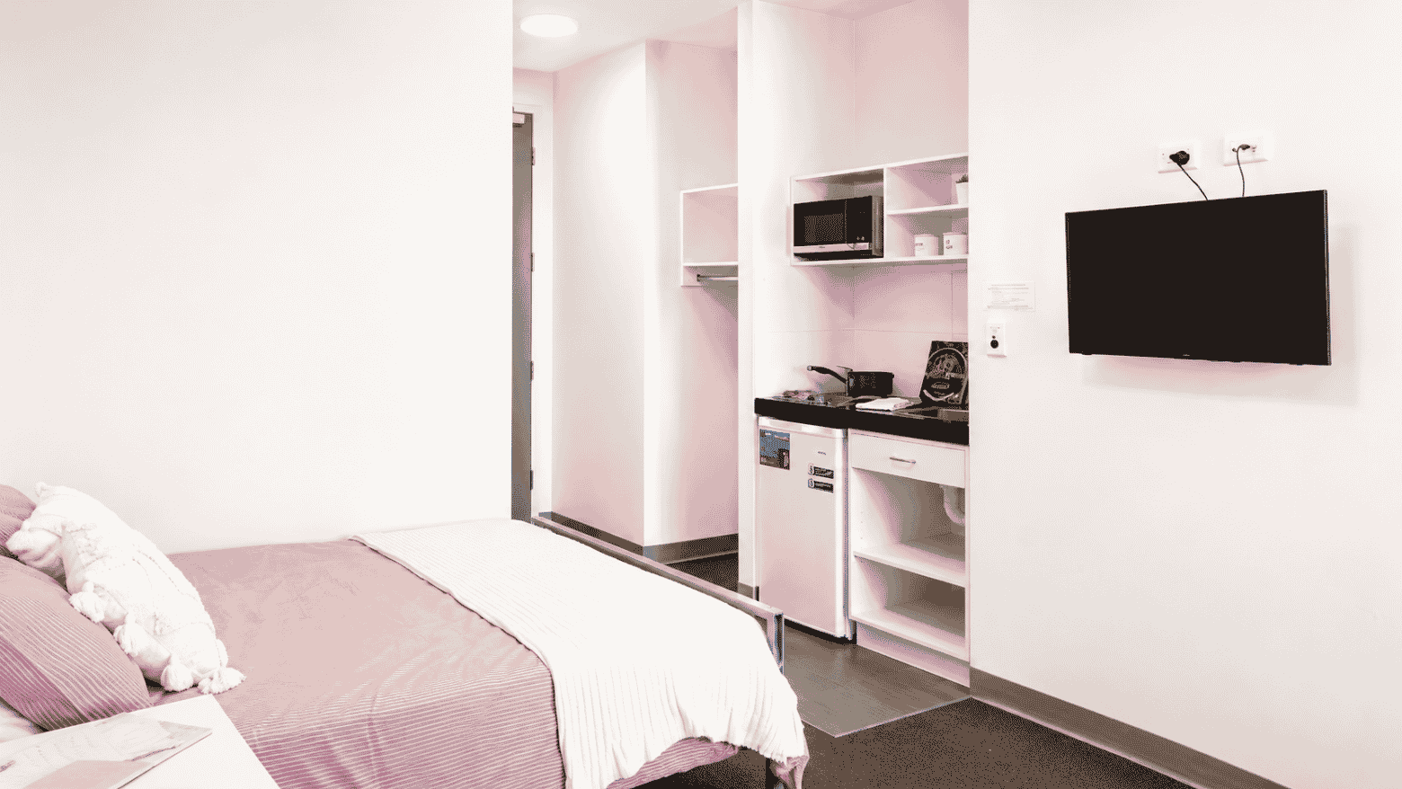 Modern student studio with pink and white decor, compact kitchenette, wall-mounted TV, and single bed at Student Living Waymouth