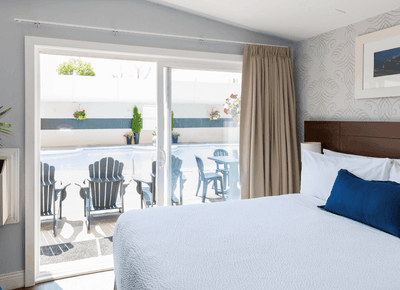 King bed beside glass patio doors opening toward the sunny outdoor swimming pool deck area with chairs at Rodd Moncton