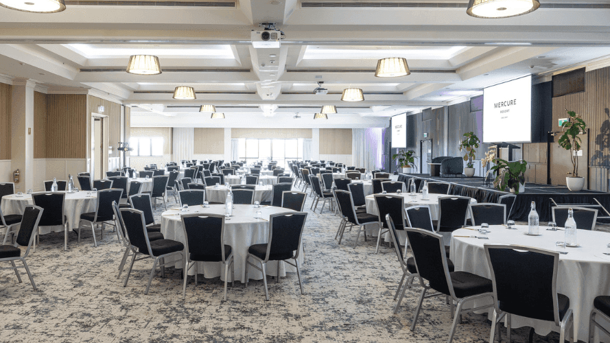 Large conference room with rows of round tables, black chairs, and a projection screen at the front.