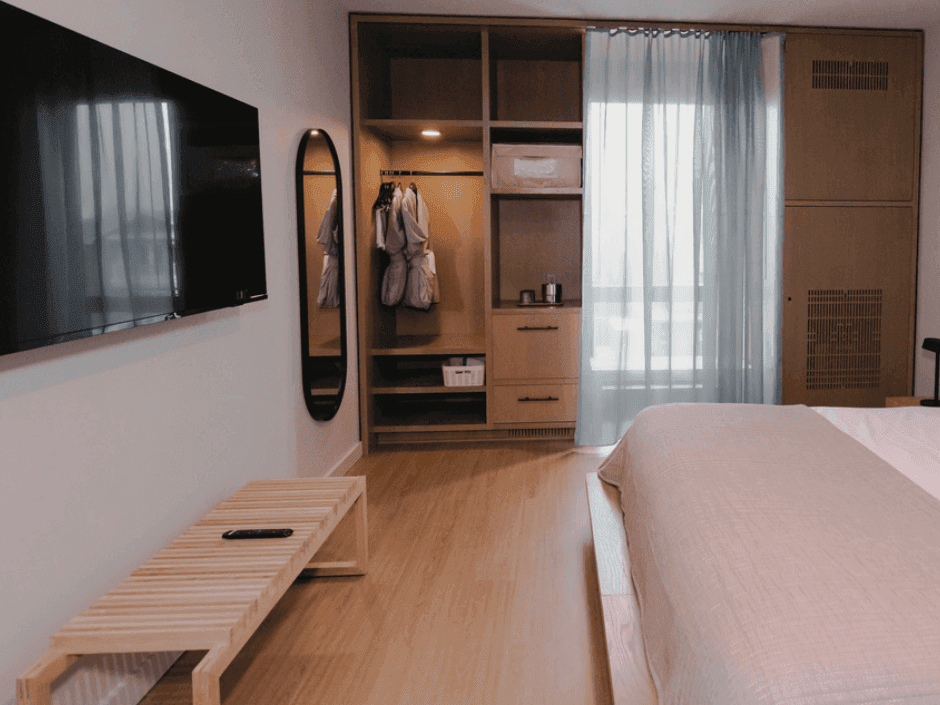 A bedroom with a bed, wooden bench, TV, mirror, and open wardrobe with clothes.
