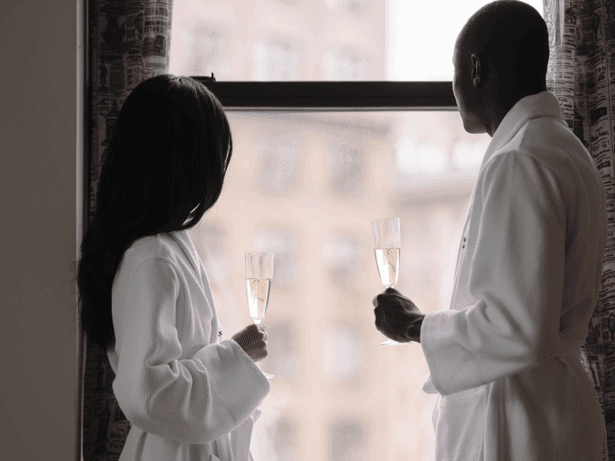 Two guests in white robes holding champagne glasses while looking out a large window with city views
