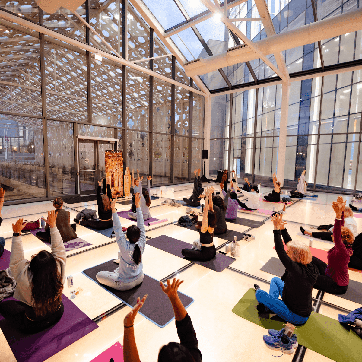 Yoga class stretching arms upward on mats under a glass atrium roof, near tall windows at Hotel X Toronto
