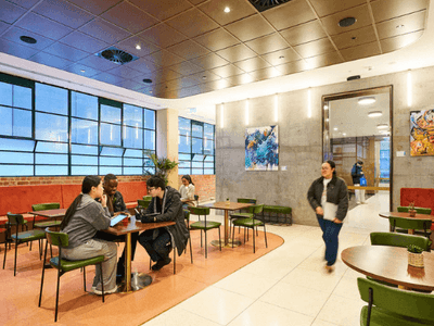 People sitting and walking in a cafe with large windows, art on the walls, and tables.