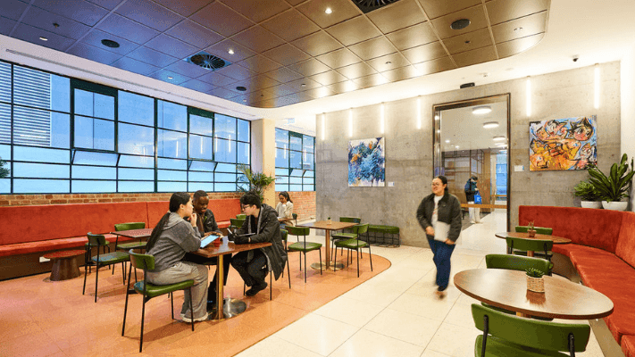 People sitting and walking in a cafe with large windows, art on the walls, and tables.