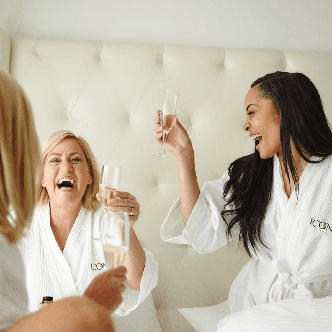 Three women in ICONA robes sitting in a bed together laughing and drinking champagne