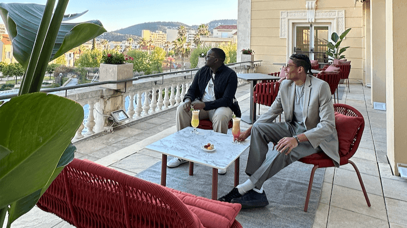 Deux hommes assis sur une terrasse, avec vue sur la ville, table basse et boissons.