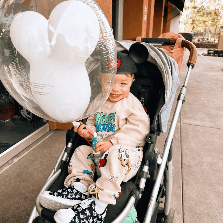 Smiling toddler wearing Mickey ears sits in a stroller holding a Mickey-shaped balloon at a theme park plaza.