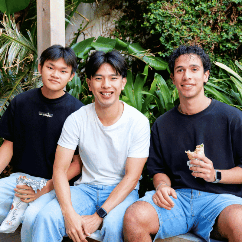 Three smiling students enjoying food together in a lush outdoor courtyard setting at UniLodge.
