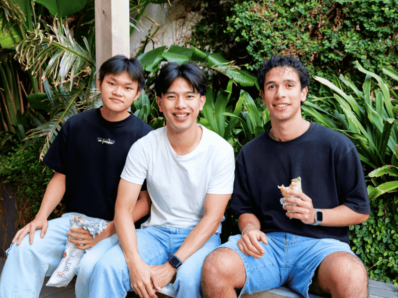Three smiling students enjoying food together in a lush outdoor courtyard setting at UniLodge.
