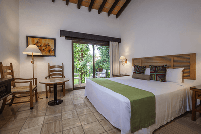 A hotel room with a large bed, wooden headboard, two chairs, a table, and a balcony with trees.