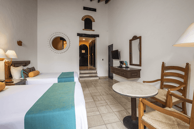 Hotel room with two beds, table, chairs, lamps, mirror, TV, and staircase in the background.