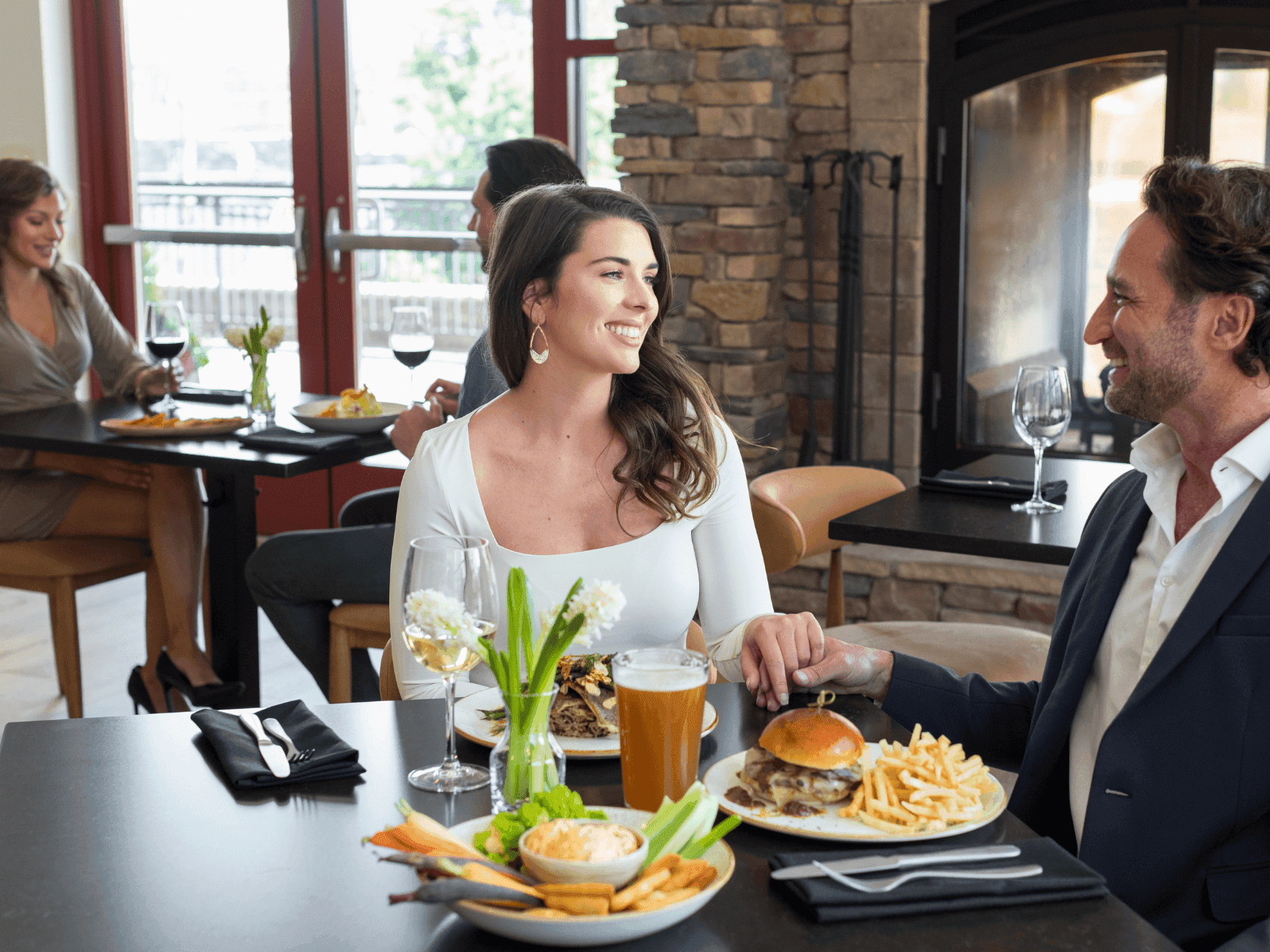 Couple dining at a restaurant, sharing a table with plates of food and drinks, surrounded by other diners.