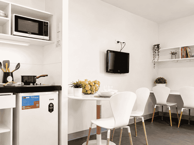 Modern student living space with compact kitchenette, dining area with white chairs, wall-mounted TV, and open shelving.