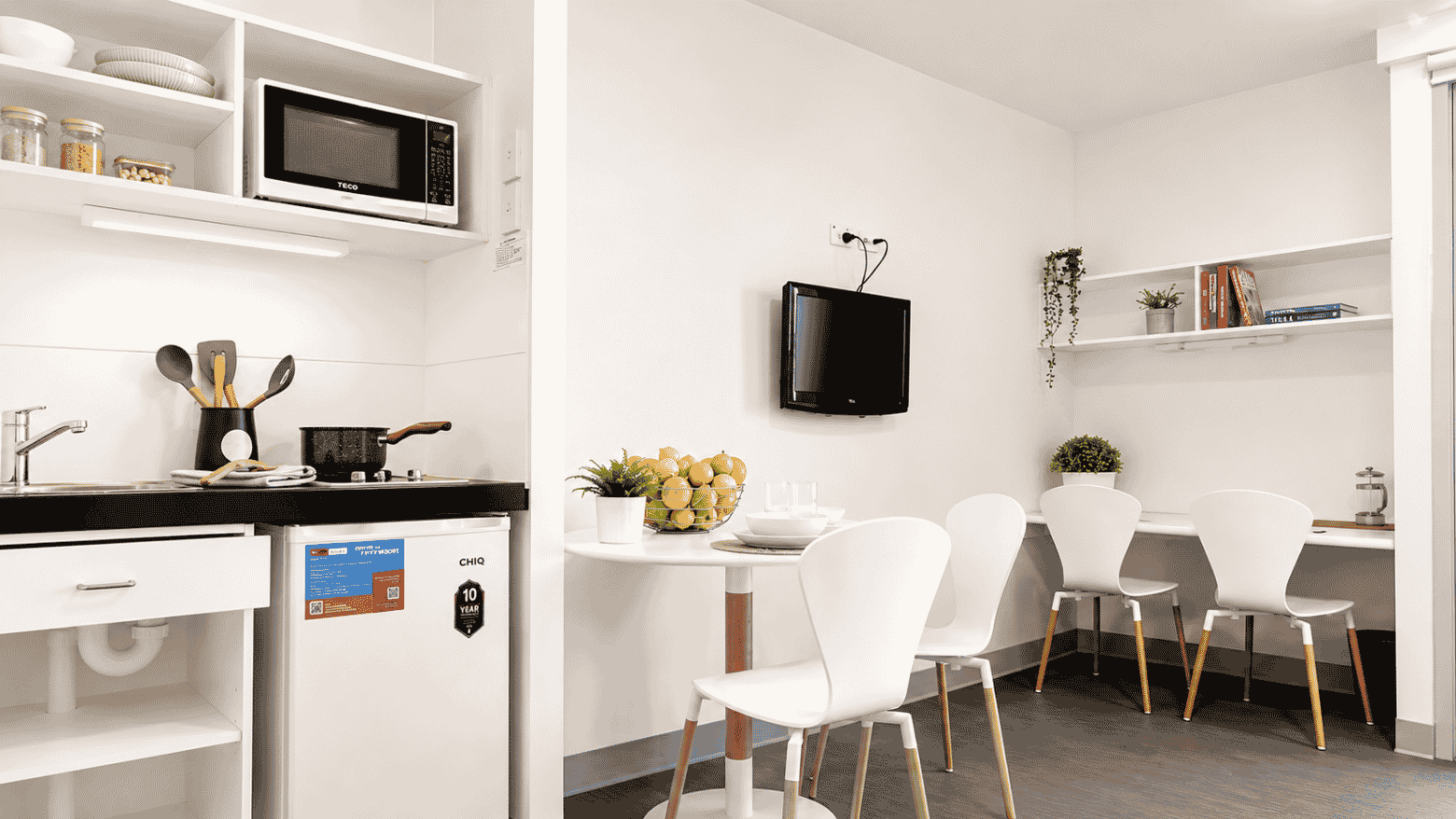 Modern student living space with compact kitchenette, dining area with white chairs, wall-mounted TV, and open shelving.