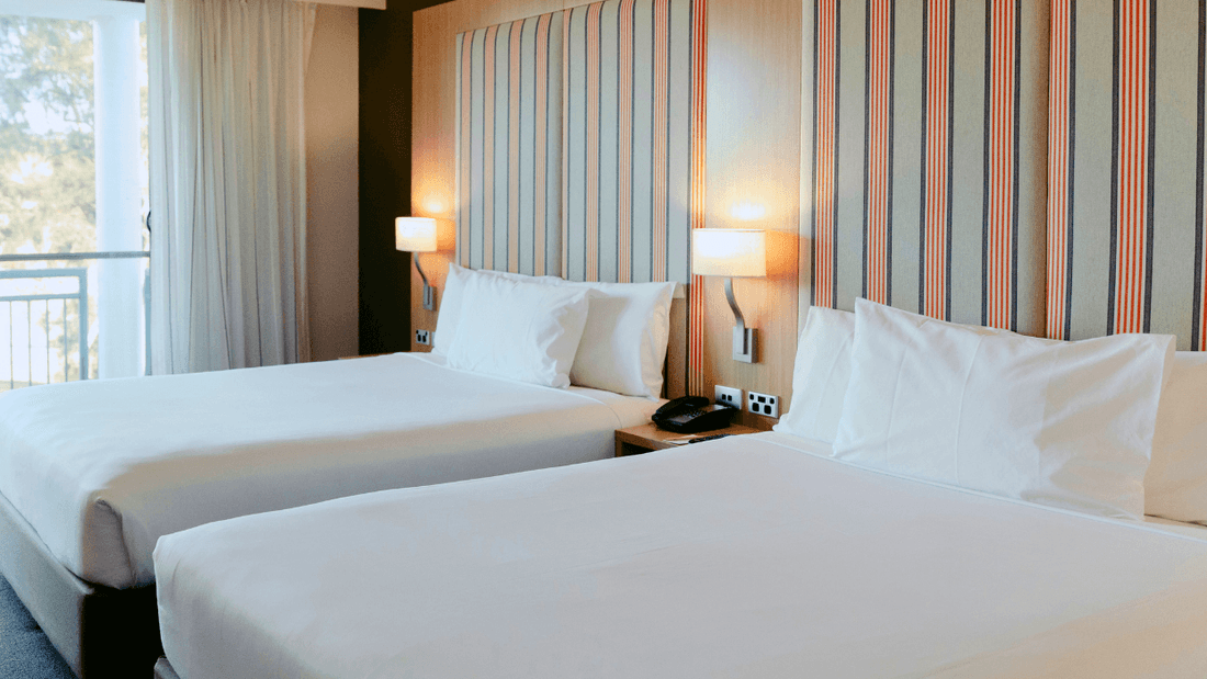 Two beds with white linens and pillows, a nightstand with lamps, and a balcony with curtains.