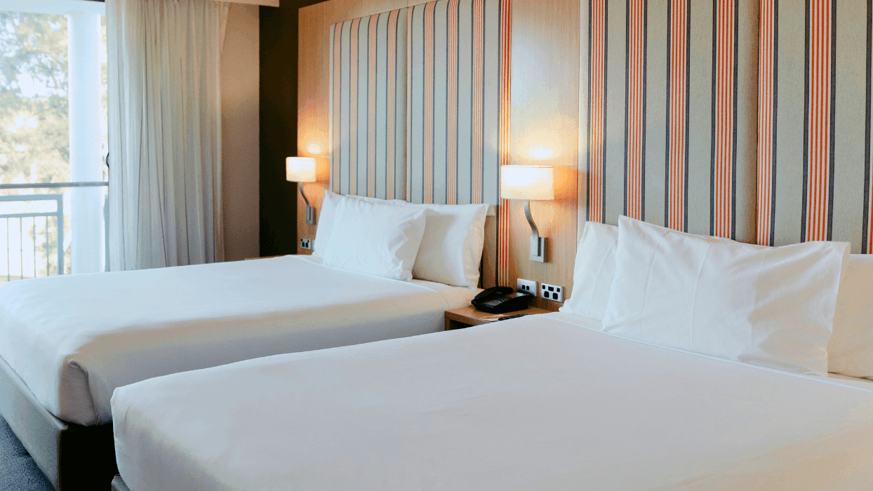 Two beds with white linens and pillows, lamps, and a nightstand in a hotel room.