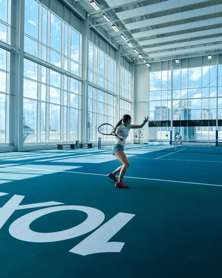 Woman playing tennis on a blue indoor court by floor-to-ceiling windows under a high ceiling at Hotel X Toronto
