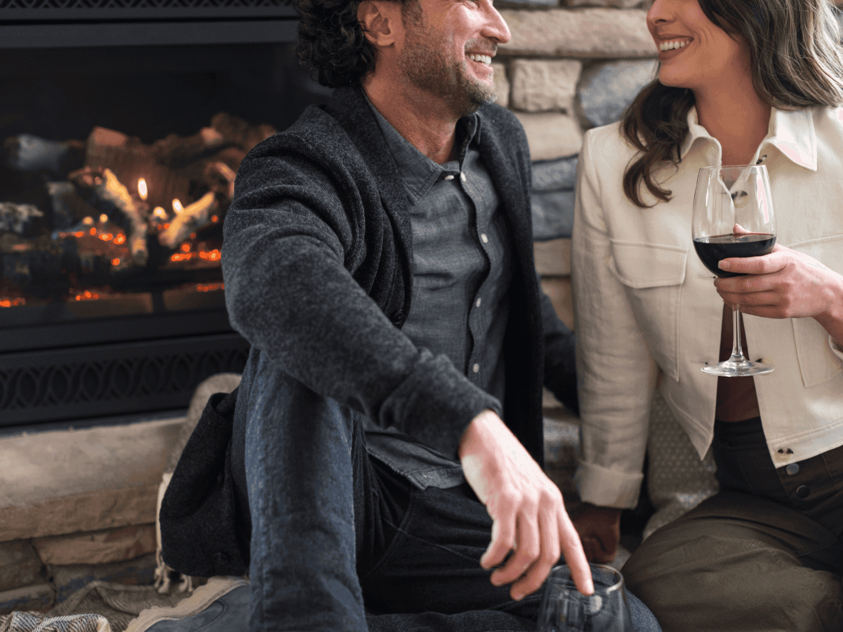 Couple sitting by a fireplace, smiling and holding wine glasses.