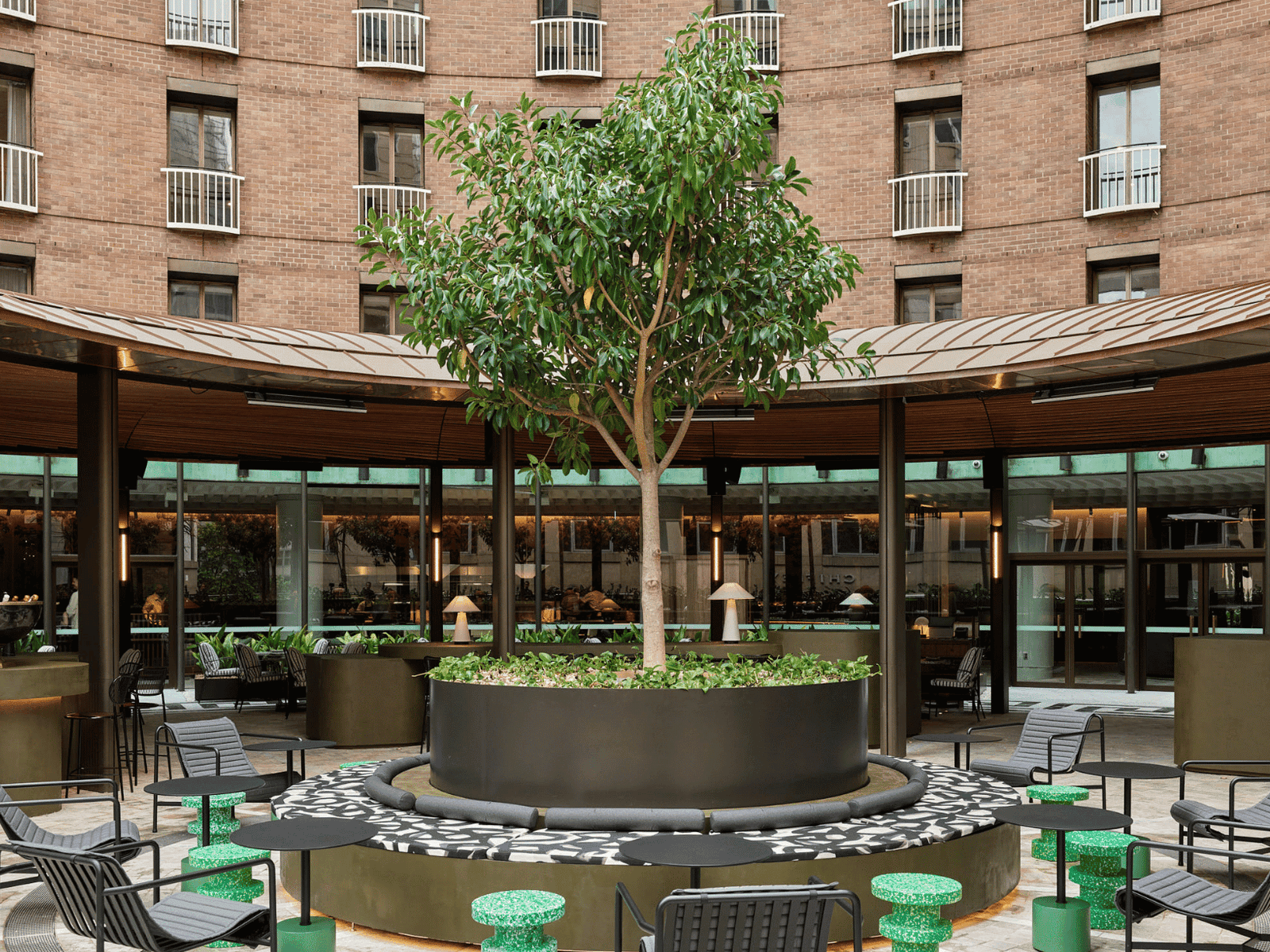 Outdoor patio of a hotel with modern furniture, a central tree, and surrounding brick buildings.