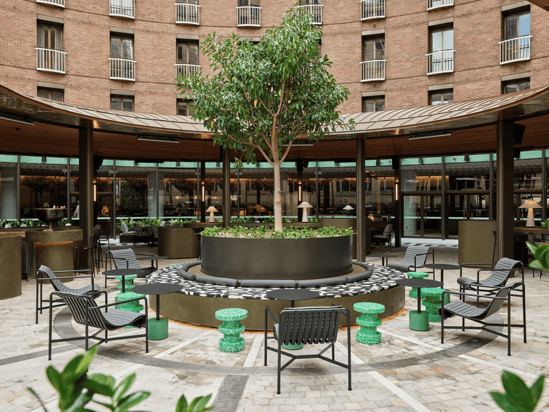 Outdoor patio of a hotel with modern furniture, a central tree, and surrounding brick buildings.