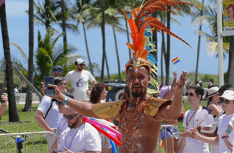 Man in orange and blue feathered headdress and gold costume dances in front of a crowd at a beach event.