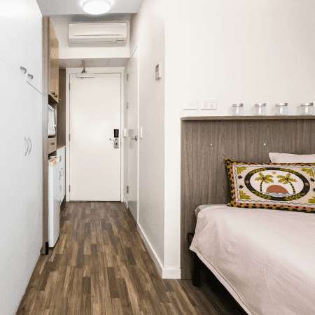 Modern hotel room with wooden floors, white walls, single bed, and white cabinets at StudentLiving Darwin.