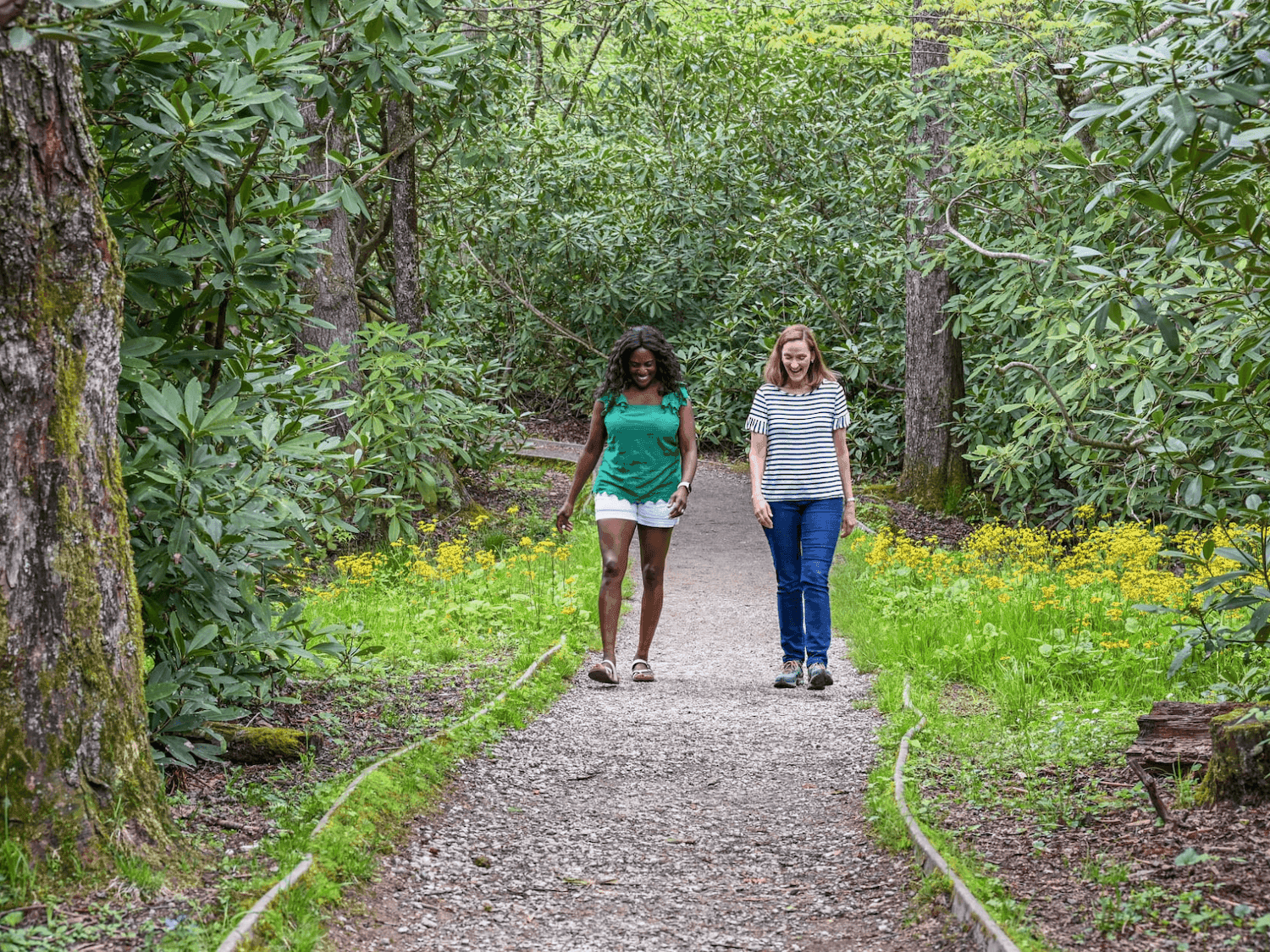 Two women walking on a gravel path surrounded by trees and greenery.
