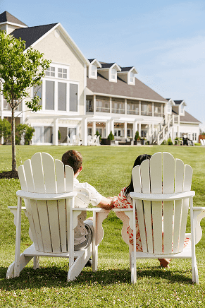 Back view of a couple sitting in white Adirondack chairs looking at the main building at Rodd Crowbush Golf & Beach Resort