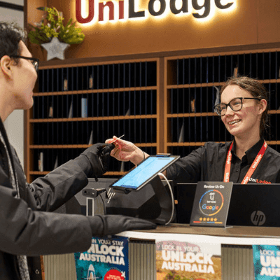 Man at front desk of UniLodge handing pen to smiling receptionist with tablet.