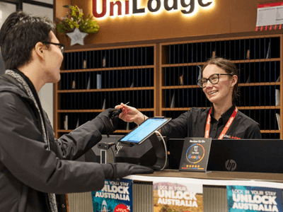 Man at front desk of UniLodge handing pen to smiling receptionist with tablet.