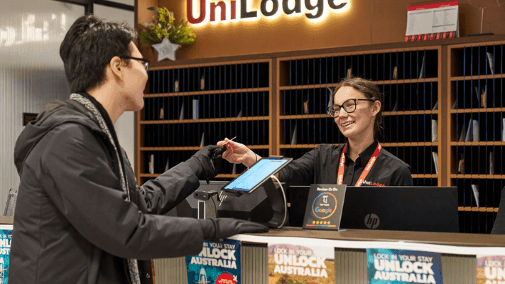 Man at front desk of UniLodge handing pen to smiling receptionist with tablet.