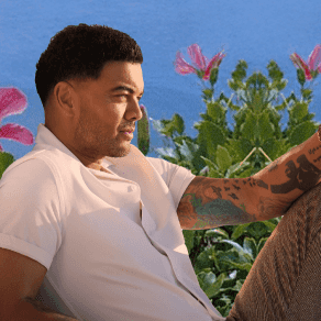 Guy Sebastian sitting with arms crossed near flowers and ocean backdrop in events module.