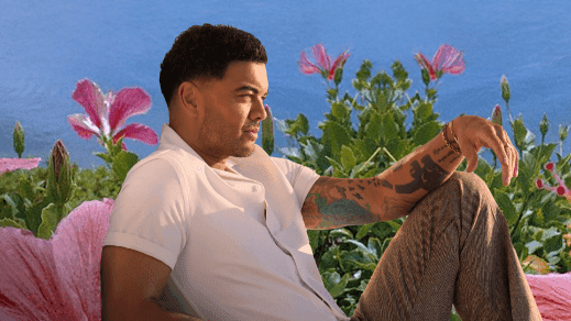 Guy Sebastian sitting with arms crossed near flowers and ocean backdrop in events module.