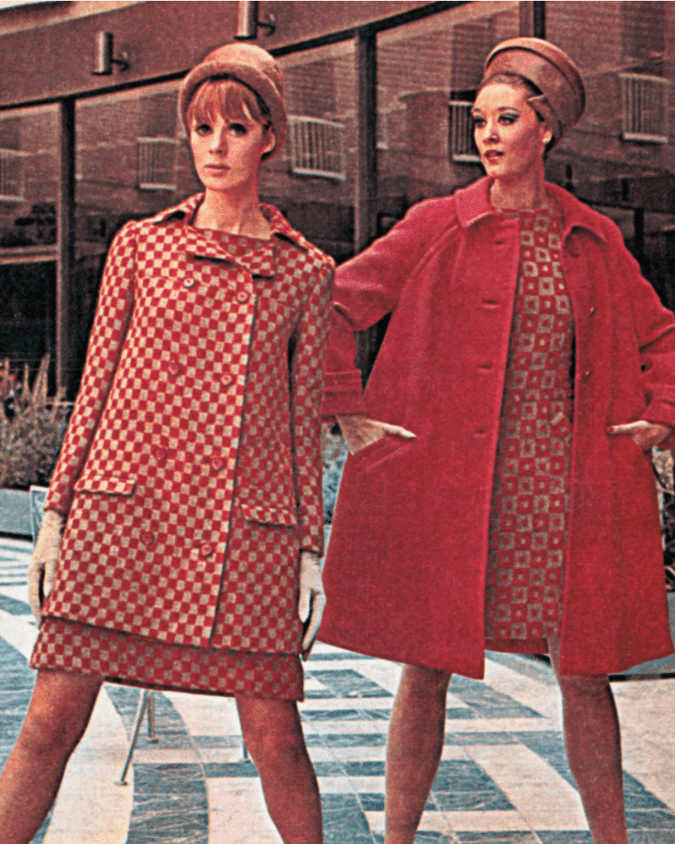 Two women in vintage red outfits and hats stand on patterned floor in front of a building.