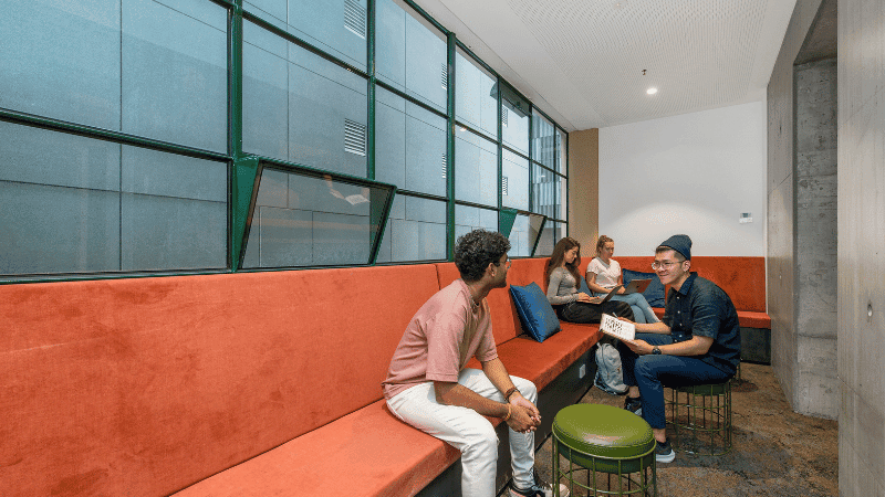 Group of people sit on red cushioned benches and stools in a modern study lounge.