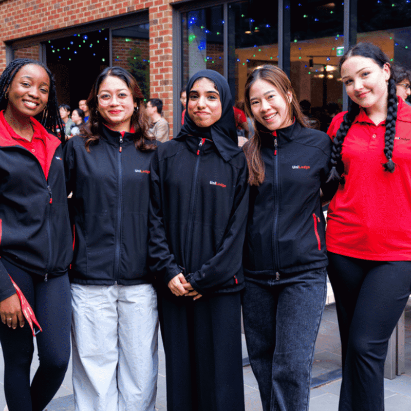 Five UniLodge staff members in branded uniforms smiling together at an outdoor courtyard event with brick building backdrop