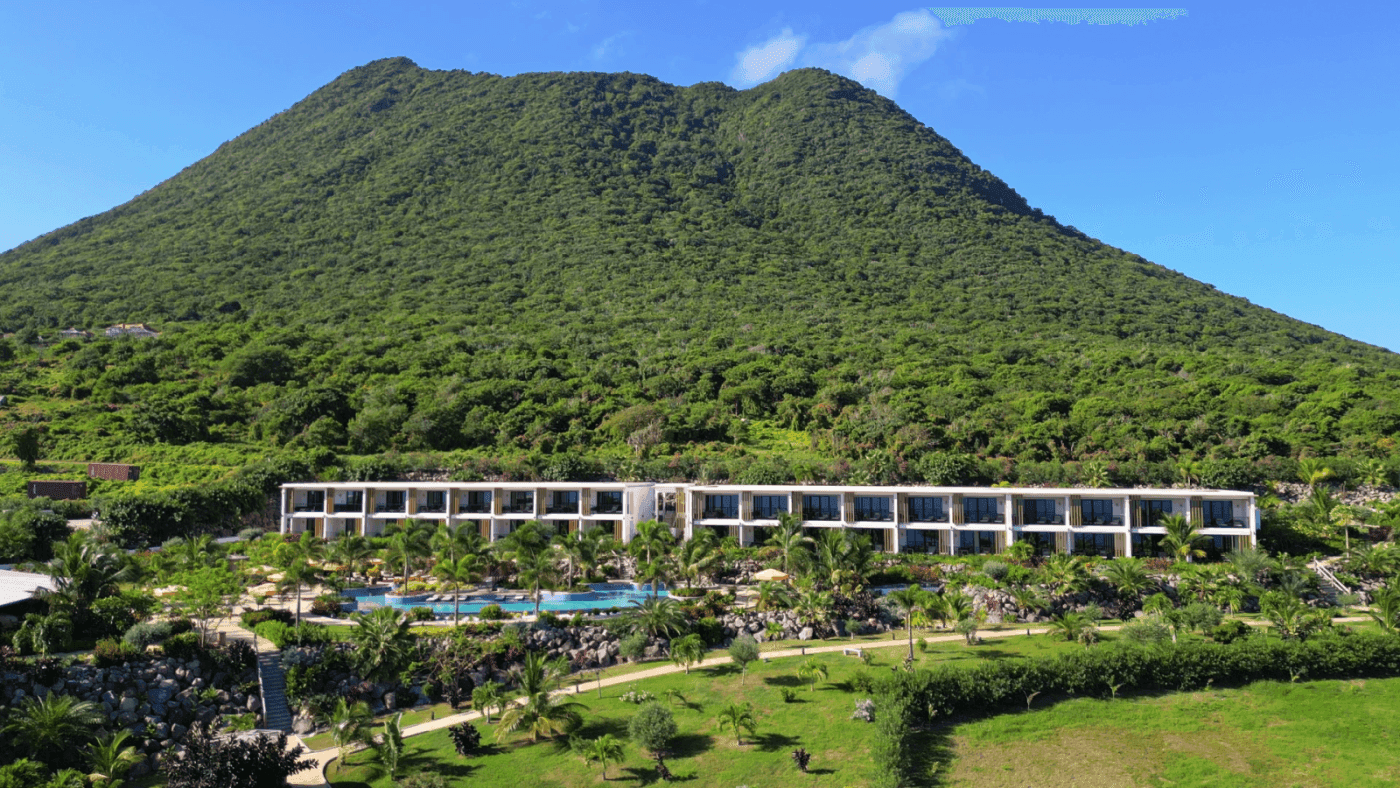 Aerial view of Golden Rock Resort surrounded by lush green mountains, palm trees & sparkling pool under a clear blue sky