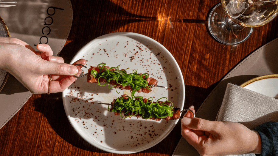 Two diners sharing an artfully plated appetizer garnished with fresh greens and spices on a wooden table.