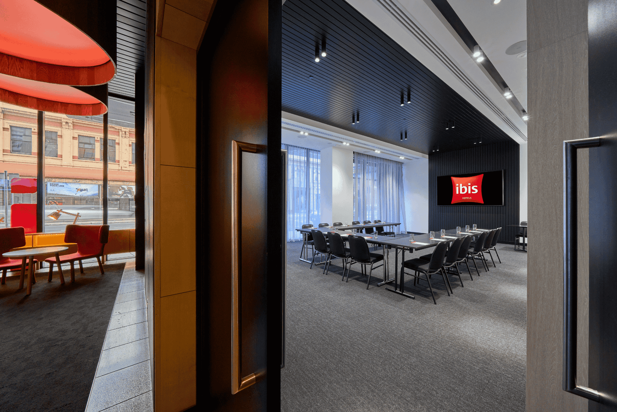 U-shaped table setup facing a wall-mounted TV in The Engine Room, alongside a cozy lounge area at Ibis Adelaide