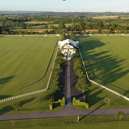 Aerial view of a paved driveway with green trees and large grass polo fields beside the main building at Polo Club Hotel