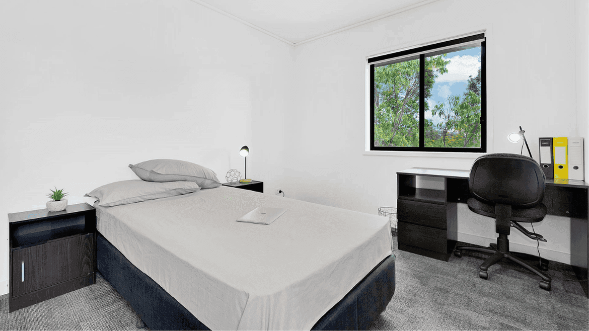 Modern bedroom with white walls, dark furniture, desk workspace, and window overlooking greenery at Springfield Central.