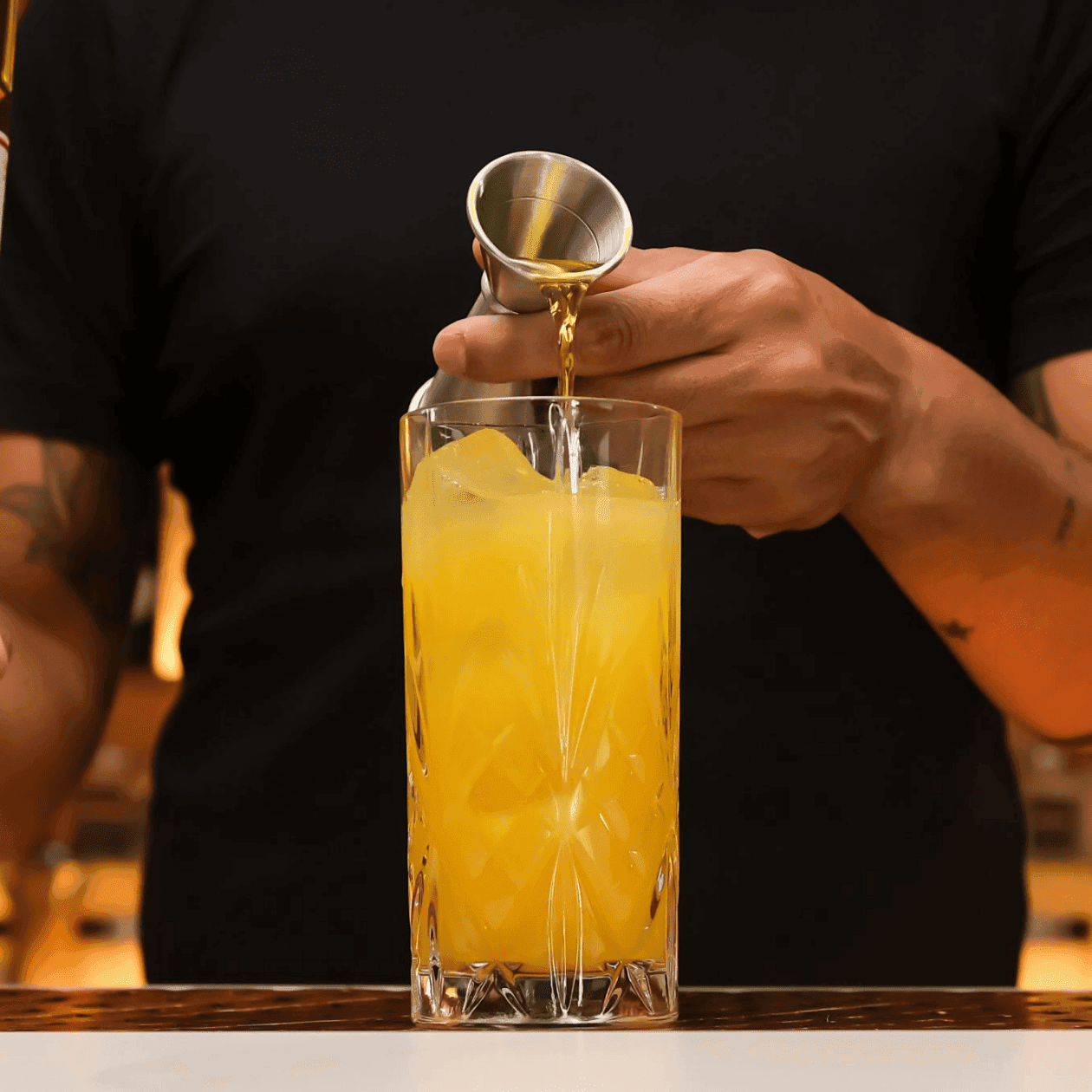 Bartender pouring orange drink from silver cocktail shaker into glass with ice, holding a bottle of G.