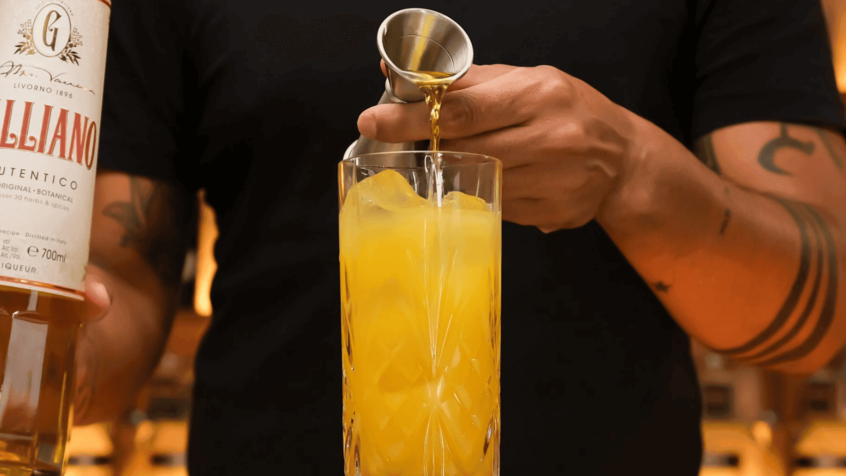 Bartender pouring orange drink from silver cocktail shaker into glass with ice, holding a bottle of G.
