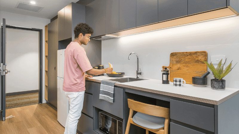 A man in a pink shirt stands at a kitchen counter preparing food near a sink.