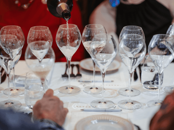 Champagne being poured into a row of glasses at a dining table, promoting FIRMA Italian’s Collector’s Champagne Table featuring five distinguished cuvées and a five-course paired dinner.