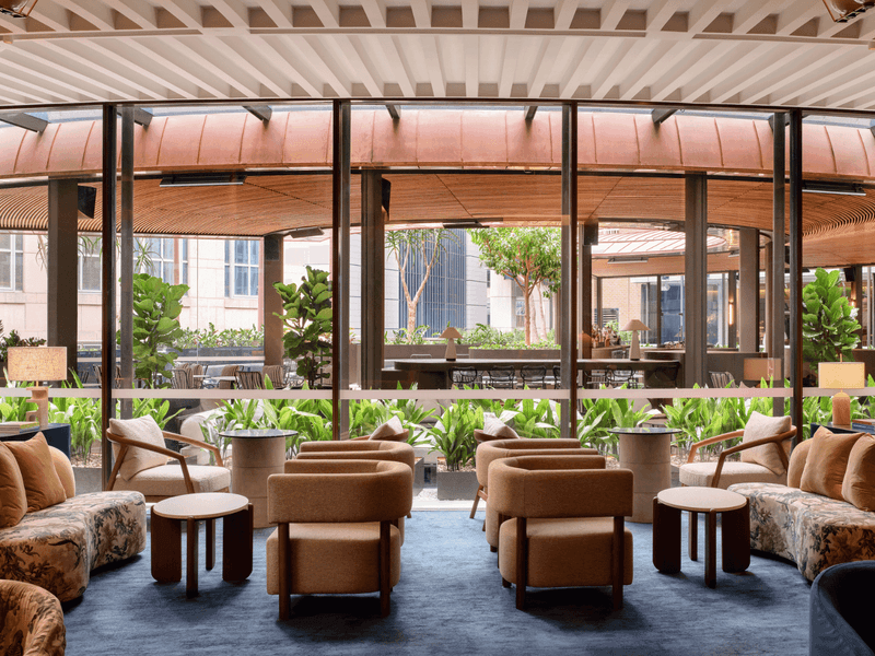 Lounge area with couches, chairs, and tables, large glass walls showing outdoor seating and greenery.