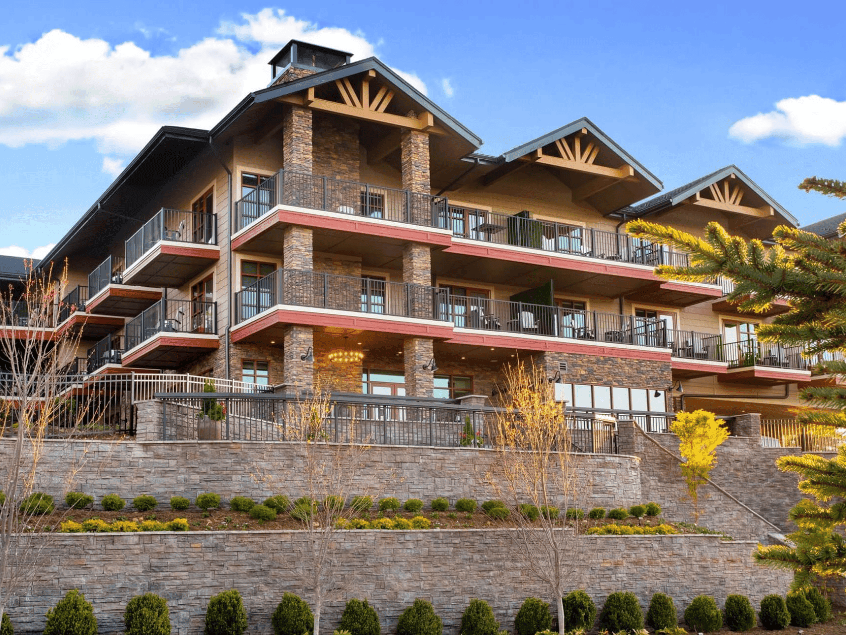 The Embers Hotel exterior with a stone facade, wood balconies and terraces, framed by pine trees and tiered gardens